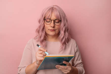 Horizontal shot of dreamy businesswoman teacher writing with pen making notes into notebook for future on clipboard isolated over pink studio background.の写真素材