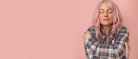 Young pretty positive caucasian woman hugging self, feeling in love and closes eyes against pink background.の写真素材