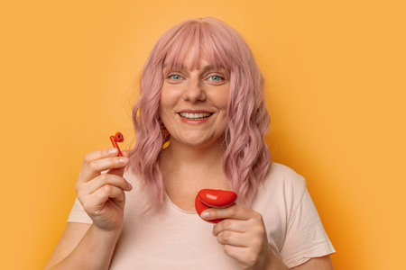 Portrait of charming caucasian woman with pink curly hair inserting wireless earphone in ear posing...