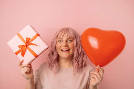 Happy positive young caucasian woman showing gift box and red heart balloon, while looking at camera isolated on pink background. Valentines Day conceptの写真素材