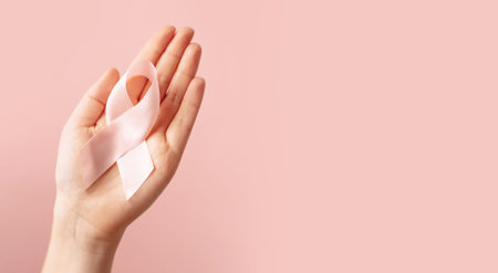 Woman hand holding pink ribbon in palm symbol of breast cancer awareness on isolated pastel pink...