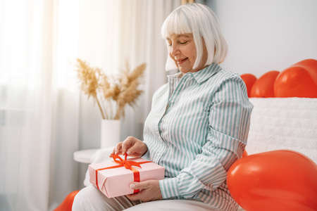 Elderly blonde haired woman 40s 50s years old wearing stylish blouse open on gift box present with red ribbon bow at home. Celebrating Valentines Day. Mother dayの写真素材