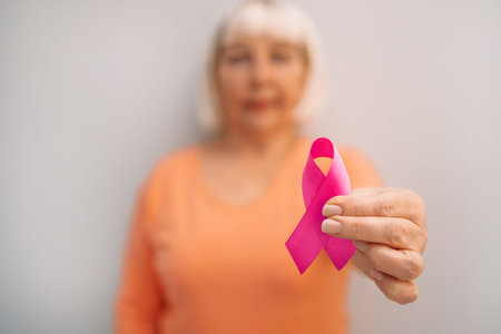 50s woman hand holding pink ribbon symbol of breast cancer awareness on isolated grey background...