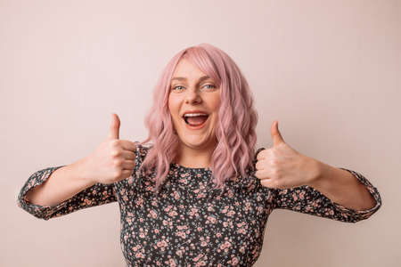 Attention promotions banner. Charming young caucasian woman with pink hair in a beautiful spring bright dress showing thumb up like gesture isolated on pastel backgroundの写真素材