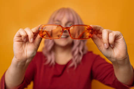 Beautiful young woman with pink hair hairstyle shows bright orange sunglasses isolated studio yellow background. Vision Careの写真素材