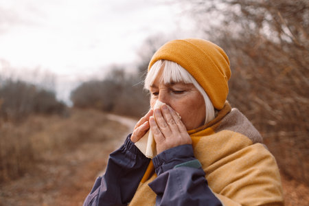 Ill young woman caught cold sneezing in paper tissue at fall city park. Sick allergic female blowing...