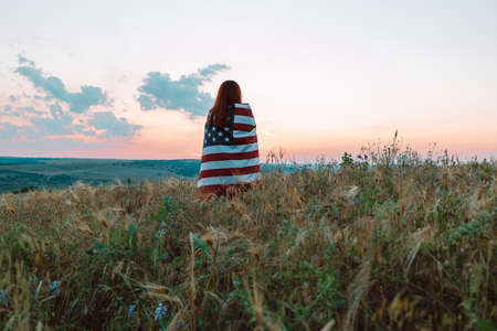 Young woman proudly hold waving american USA flag in the summer field. Patriot raise national american flag against blue sky. Independence Day, 4th July.の写真素材