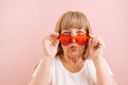 Happy beautiful smile 50s woman in red glasses in good mood sgives an air kiss isolated over pink...