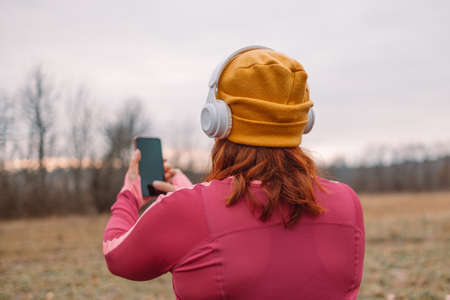 Fitness young runner with overweight in sportswear uses a smartphone listening music during training outdoors in city autumn park. Healthy lifestyle concept.の写真素材