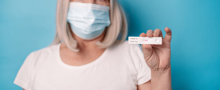 Woman in protective medical mask holding an antigen test with positive result for COVID-19 pandemic posing over bright blue color backgroundの写真素材