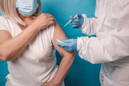 Close up of doctor giving covid-19 vaccination against covoravirus to elderly 50s woman, wearing face mask over blue background.の写真素材