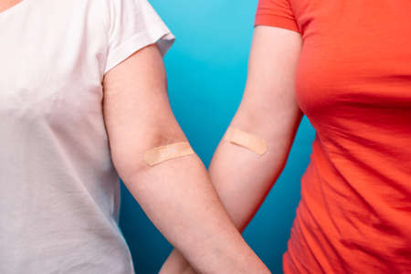 Two woman in T-shirts with hands taped patch after giving blood on a blue background. World blood donor day and save life concept.の写真素材