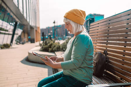 50s 60s Woman using digital tablet PC sitting on a modern wood bench in city walkingの写真素材