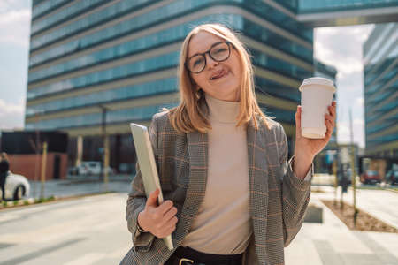 Caucasian blonde 25s business woman in trendy clothes with a laptop and take away paper coffee cup, shows tongue, posing outside near city business centreの写真素材