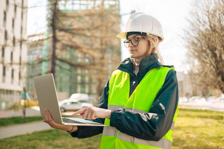 Industrial engineer woman in working uniform connected with PC laptop standing at the construction...