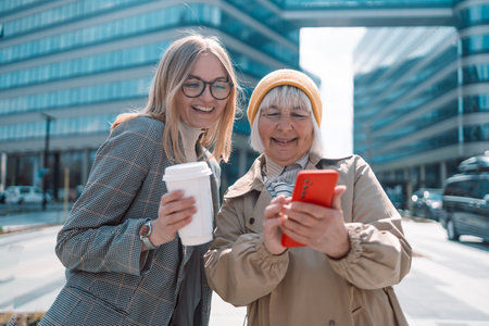 Two young caucasian women enjoy social media on their smart phones during their lunch break at their company court yard, modern office building in the background, good for modern lifestyle.の写真素材