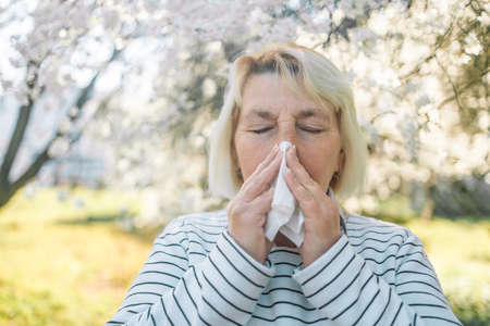 50s woman sneezing and wipes nose with napkin during walking in spring park. Flu season, cold rhinitis. Allergic people. Sick person.の写真素材