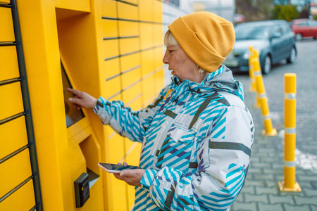 Caucasian50s 60s woman putting parcel to cell of automatic post terminal and showing smartphone with...