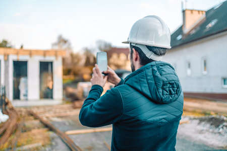 30s 40s bearded engineer worker with helmet on head standing outside of building in construction process, taking photo on smart phoneの写真素材