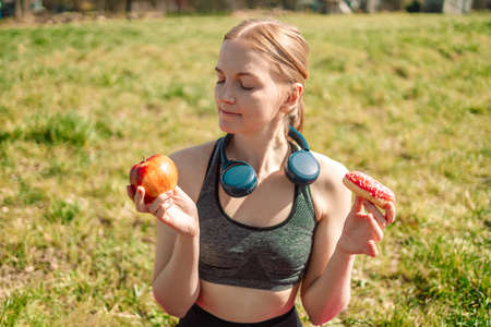 Woman making choice between apple and donut. Dieting concept. Blonde girl holds a pink donut and apple. Sweets are unhealthy junk food. Dieting. Healthy Eating.の写真素材