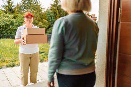 Delivery woman carry two paper post boxes deliver to customer in front of door at home. Express delivery service, online shopping and logistics concept.の写真素材