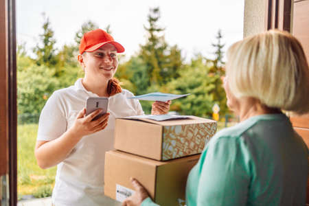 Young delivery service woman in uniform with woman customer receiving parcel post paper box and mail bill from courier at home, cargo shipping. Express delivery service, online shopping and logistics concept.の写真素材