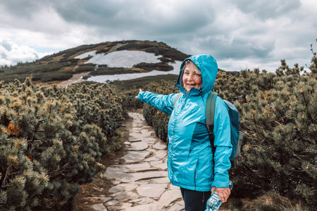 Happy senior caucasian 60s blonde woman in sportswear shows the direction to the top mountains. Beautiful Mountain nature with forest plantsの写真素材