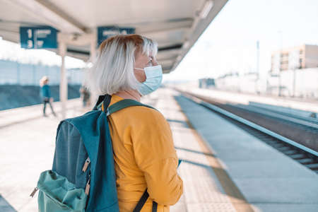 Senior 50s female traveler waiting with a blue backpack at the modern train transport stop platform outdoors, back view.の写真素材