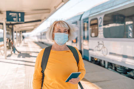 Portrait of Caucasian young woman traveller new normal using smartphone wear face mask waiting at train platform. Social distancing.の写真素材