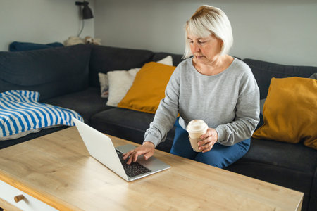 Beautiful senior lady with blonde hair work at the notebook sit down on the sofa at home. Technology...