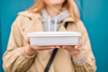 Woman hands holding empty plastic disposable food container mock up with copy space on blue background. Delivery food concept.の写真素材