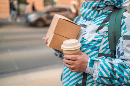 Stack of food delivery boxes. Anonymous hands holding stack of delivery boxes and coffee cup outdoors.Craft packaging of delivered food in hands.の写真素材