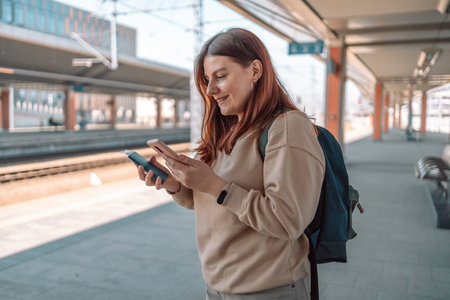Young redhead woman with passport waiting on station platform and using smart phone. Railroad...
