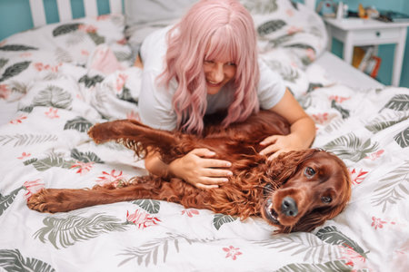 Happy smiling woman with pink hair plays with Irish Setter dog in bed early in the morning.