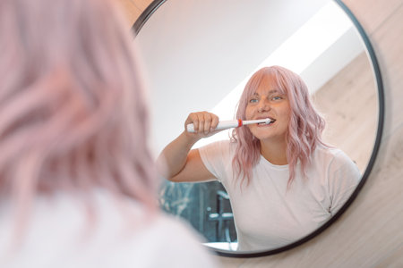Caucasian pink hair woman brushing teeth with electric toothbrush in bathroom. Modern domestic lifestyle. Hygiene and dental careの写真素材