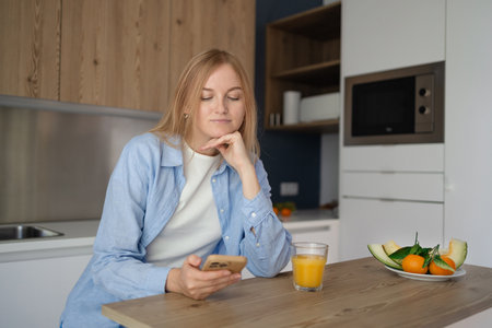 Meditative young blonde woman using smartphone and drinking orange juice while sitting at the kitchen, girl is messaging online on the phone in the morning.の写真素材
