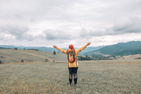 A woman with a backpack stands on the top of a mountain with her hands raised and admires the beauty...