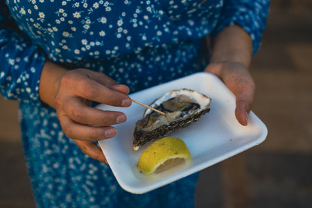 Close up female hand holding take away food tray with fresh opened oyster and lemon slices at street food market, festival, event. Selective focus. Valencia, Spain, Europe.の写真素材
