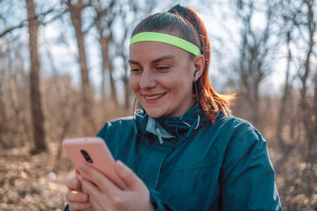 Social media and sport. Young smiling girl making sport and running in the park using her phone to listen the music with wireless headphones in the city watching the screen at autumn parkの写真素材