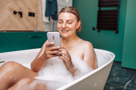 Young smile woman taking a bath and talking on phone, lying in bathtub with foam at home.の写真素材