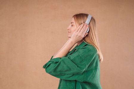 Pretty young woman girl in a stylish green jacket posing isolated on pastel wall background studio portrait. People emotions lifestyle concept. Mock up copy space. Listen music with headphones.の写真素材