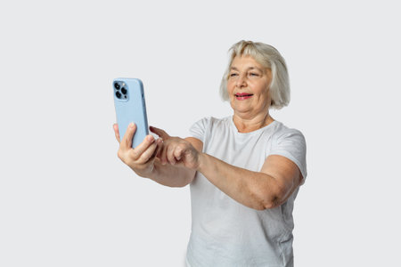 Vivid elderly blonde woman lady 40s years old wears white t-shirt doing selfie shot on mobile cell phone post photo on social network isolated on grey background studio portraitの写真素材
