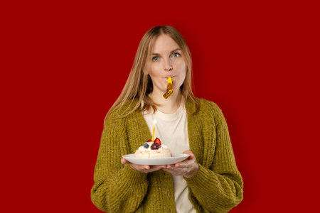 Holidays and celebration. Excited woman celebrating birthday, blowing candle on cake, wearing party cake and having fun, standing over red background.の写真素材