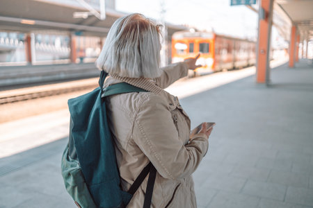 Old happy woman with backpack in yellow hat checking her ticket shows the direction of the path on background trainの写真素材