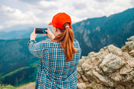 Young woman is standing on the summit of a mountain, taking a photo with a camera. Woman is positioned at the very top of the mountain, with a breathtaking scenic view behind her.の写真素材