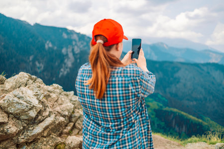 Young woman relaxing on the top mountains, holds a mobile phone in her hands and takes pictures of the landscape. Back view.の写真素材