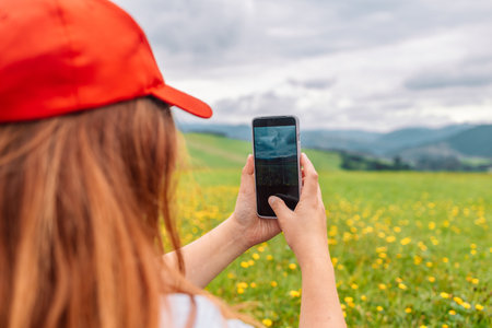 Young cheerful woman traveler in a jacket with a backpack holds a smartphone in her hands while standing on the shore of the mountainの写真素材
