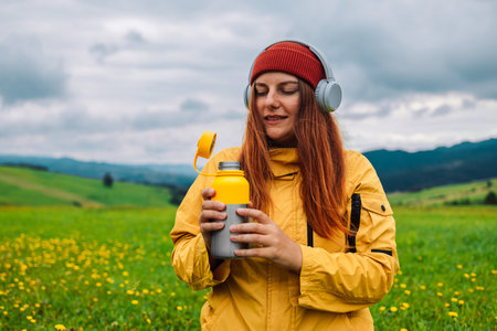 Pretty young girl traveling sitting in mountains drinking tea from camping flask enjoying trip,...