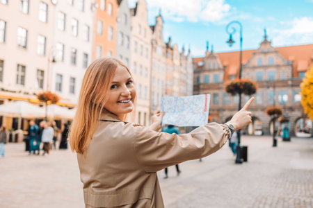 Back view of lost young female tourist holding a paper map on central square in Gdansk old town. Traveling Europe in autumn. High quality photoの写真素材