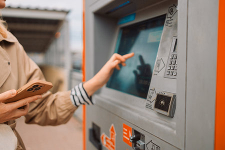 A side view of a young beautiful elegant blonde woman paying for service underground parking or buying a subway or train ticket using an electronic self-service kiosk. Travel, airport and couple at check in machine with passport and ticket for flight.の写真素材